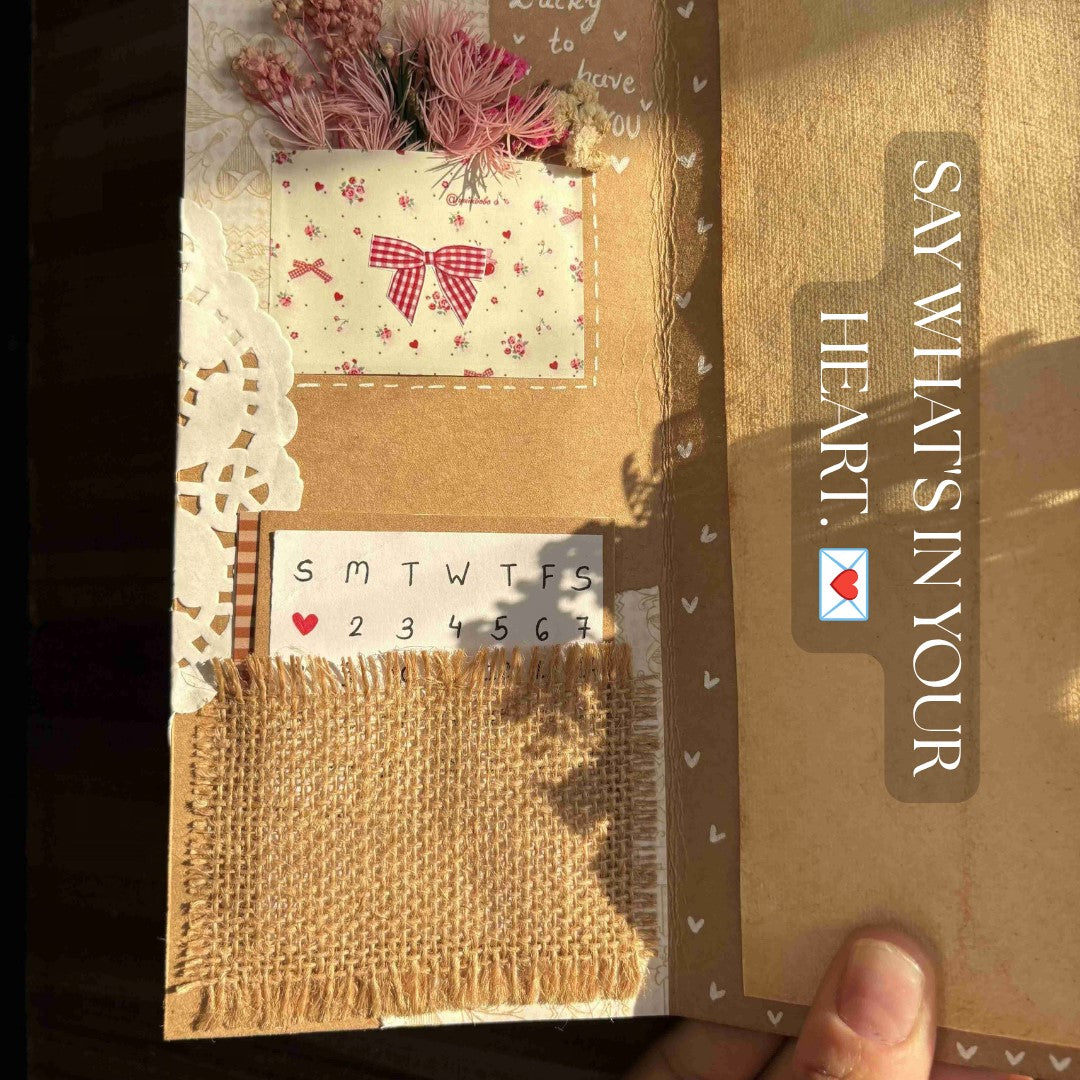 A handmade greeting card or scrapbook album page with a rustic design, featuring burlap texture, a red-and-white bow patch, dried pink flowers, a small calendar label, and vertical text reading 'SAY WHAT'S IN YOUR HEART.'